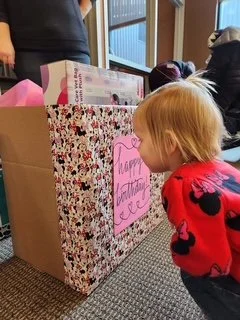 Child with blonde hair wearing a red sweater with black hearts, leaning in to look at a large, decorated gift bag with a pink note saying "Happy Birthday" in a room with other people.