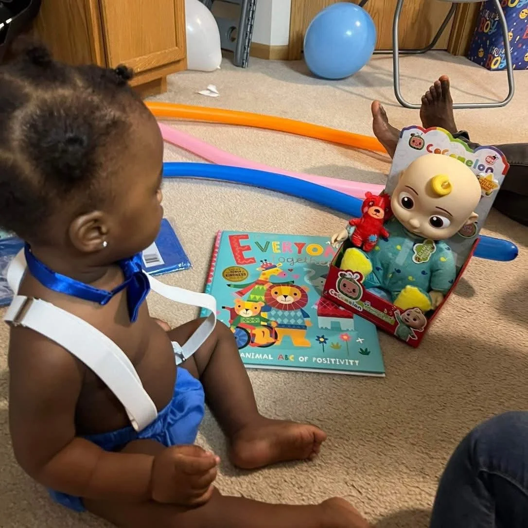 Young baby with braided hair and a blue bow tie seated on a beige carpet, facing a boxed baby doll toy with a yellow curl on its head and a red teddy bear, surrounded by colorful balloons, a book titled 'Every Animal' with illustrations of lions and 