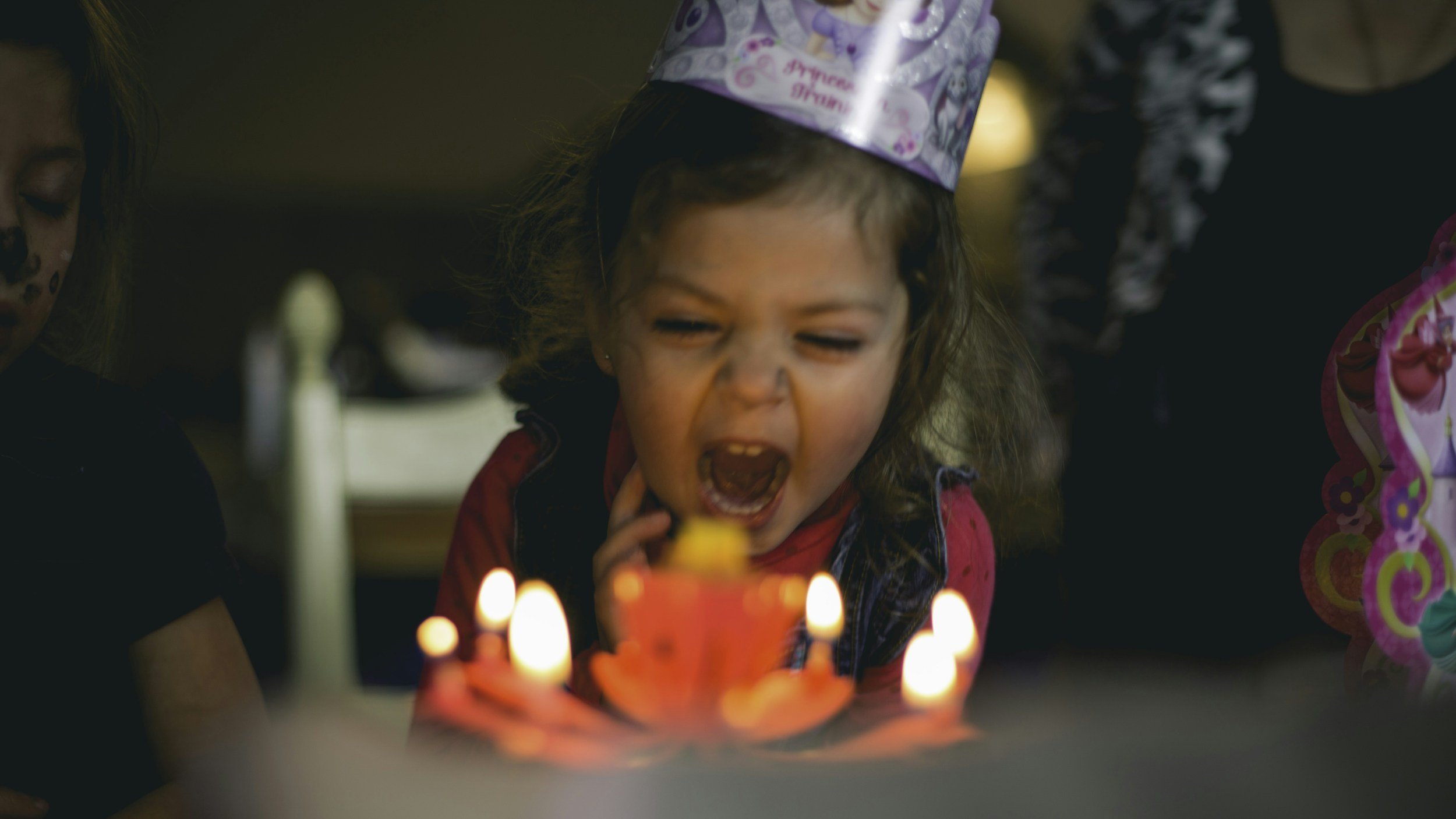 Young girl wearing a birthday hat blowing out candles on a birthday cake surrounded by lit candles.