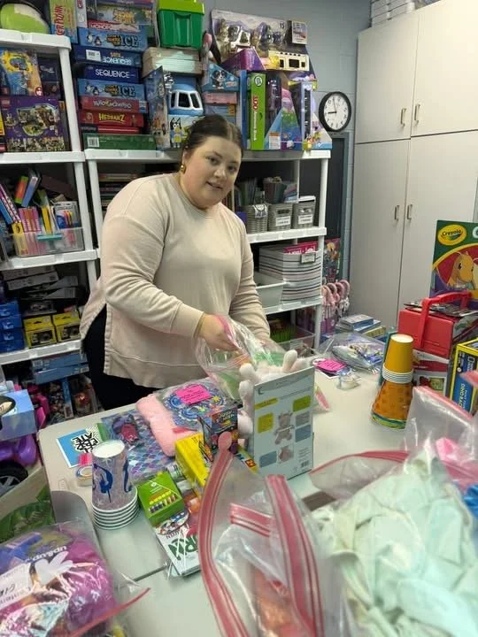 A woman in a beige sweater and gloves arranges items on a table surrounded by toys and supplies in a store or classroom setting.