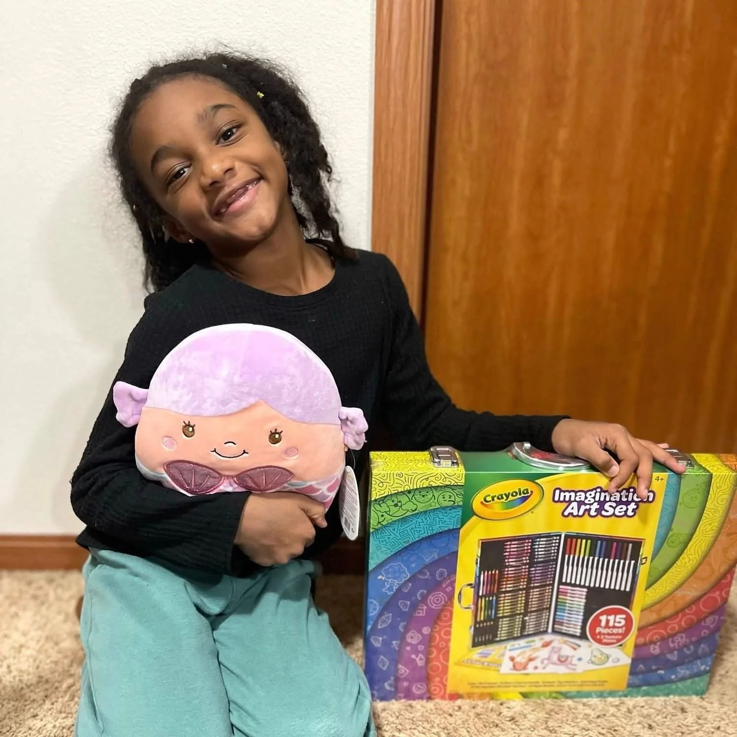 Young girl with a smile holding a plush toy and a Crayola imagination art set box, sitting on carpeted floor with wooden door in background.