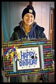 Smiling woman in a hoodie and beanie holding a birthday cake with colorful wrapping and a 'HAPPY BIRTHDAY' sign.