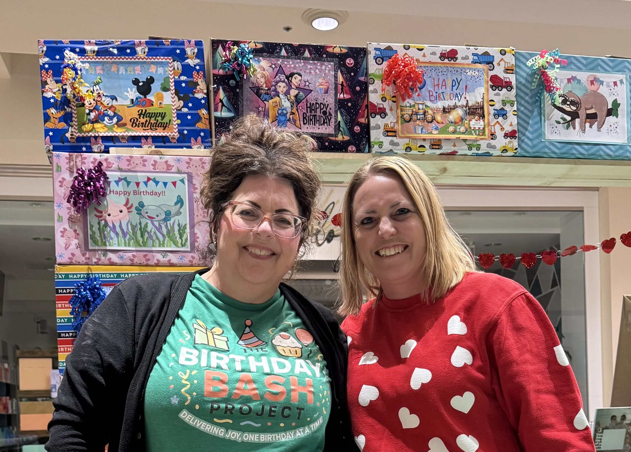 Two women smiling at a birthday party with colorful birthday-themed boxes and decorations in the background.