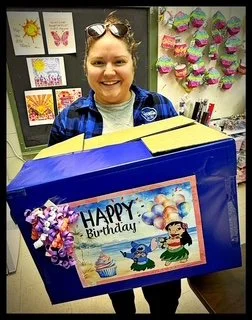 A woman holding a large birthday card box with a colorful 'Happy Birthday' design, standing in a decorated room.
