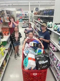 A group of children shopping for sports equipment inside a store, with a woman supervising. One child pushes a shopping cart filled with tennis rackets, while others look at products on the shelves.