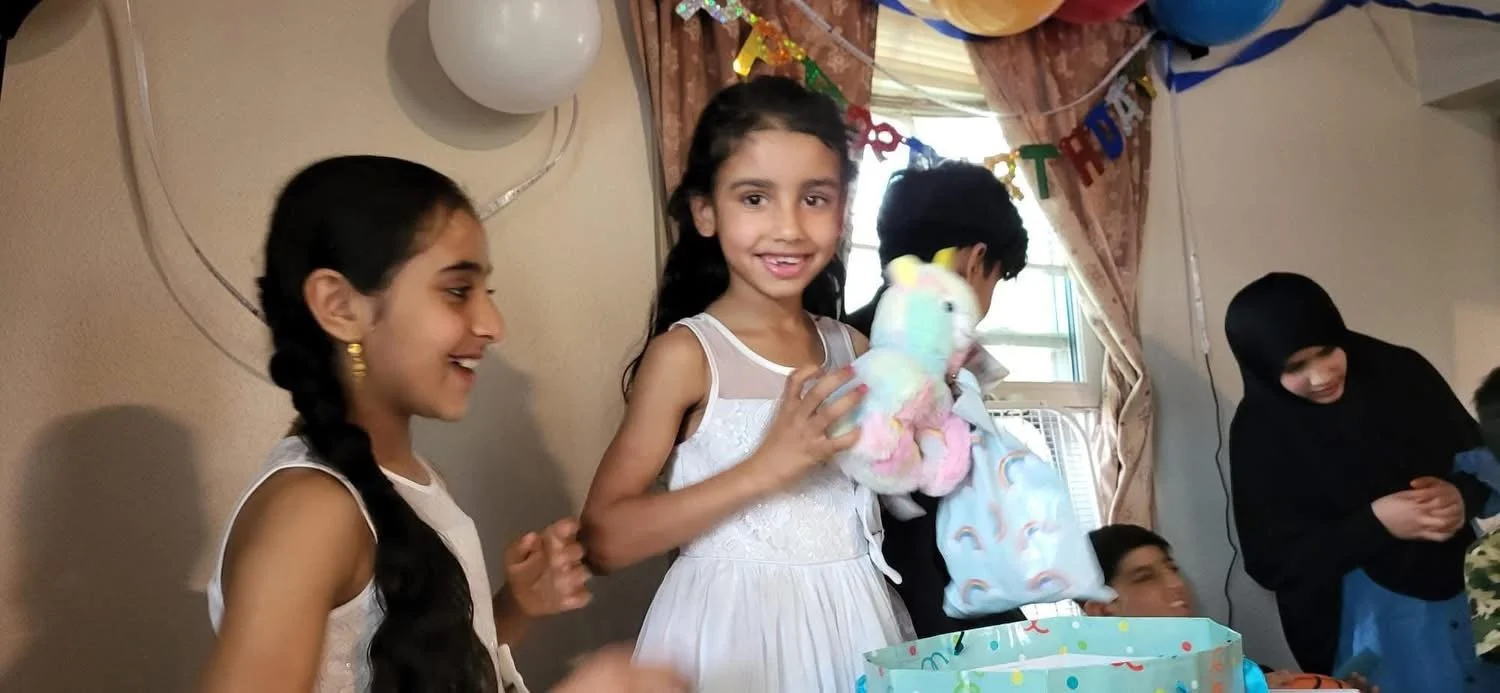 Group of children celebrating birthday party indoors, decorated with colorful banners, balloons, and a birthday gift.