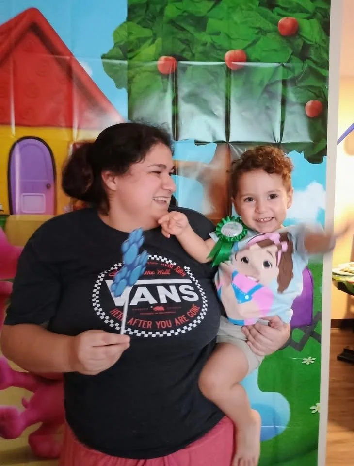A woman and a young girl smiling together, with the woman holding a balloon and the girl wearing a birthday badge, in front of a colorful cartoon backdrop.