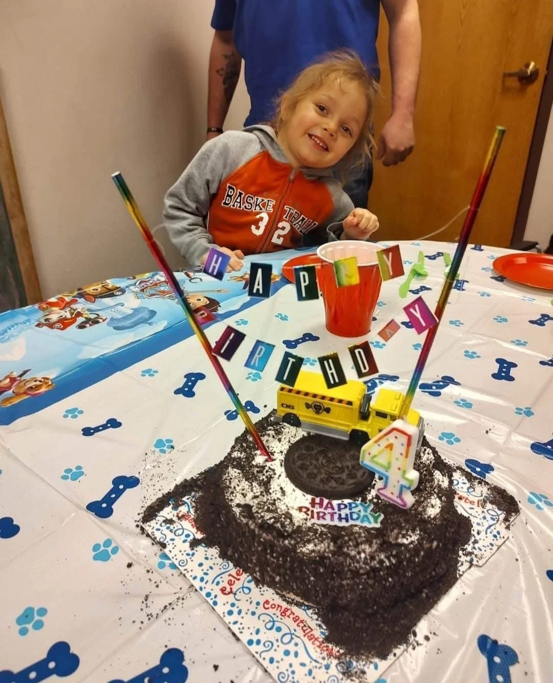 Little girl celebrating her birthday with a chocolate cake decorated with a yellow toy school bus and a '4' birthday candle, surrounded by birthday decorations, party banners, and a tablecloth with dog bone and paw prints.