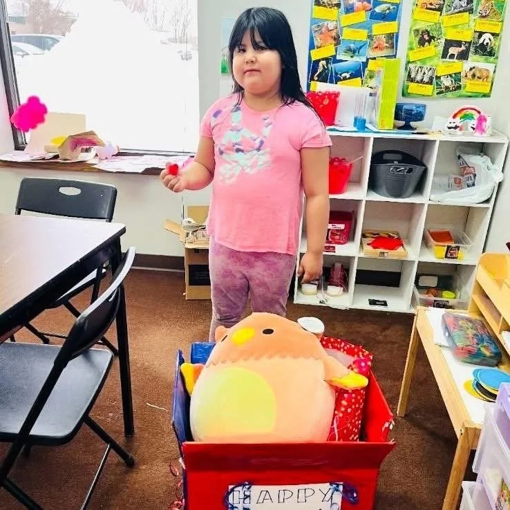 A young girl with black hair wearing a pink shirt and purple leggings standing in a classroom. She holds a small red object. In front of her is a large plush turkey stuffed animal in a red cart with a sign that reads "HAPPY". The classroom has shelve