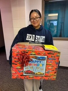 A young woman with dark hair and glasses holding a large, colorful birthday gift box with a 'Happy Birthday' label, indoors near a window and tiled wall.