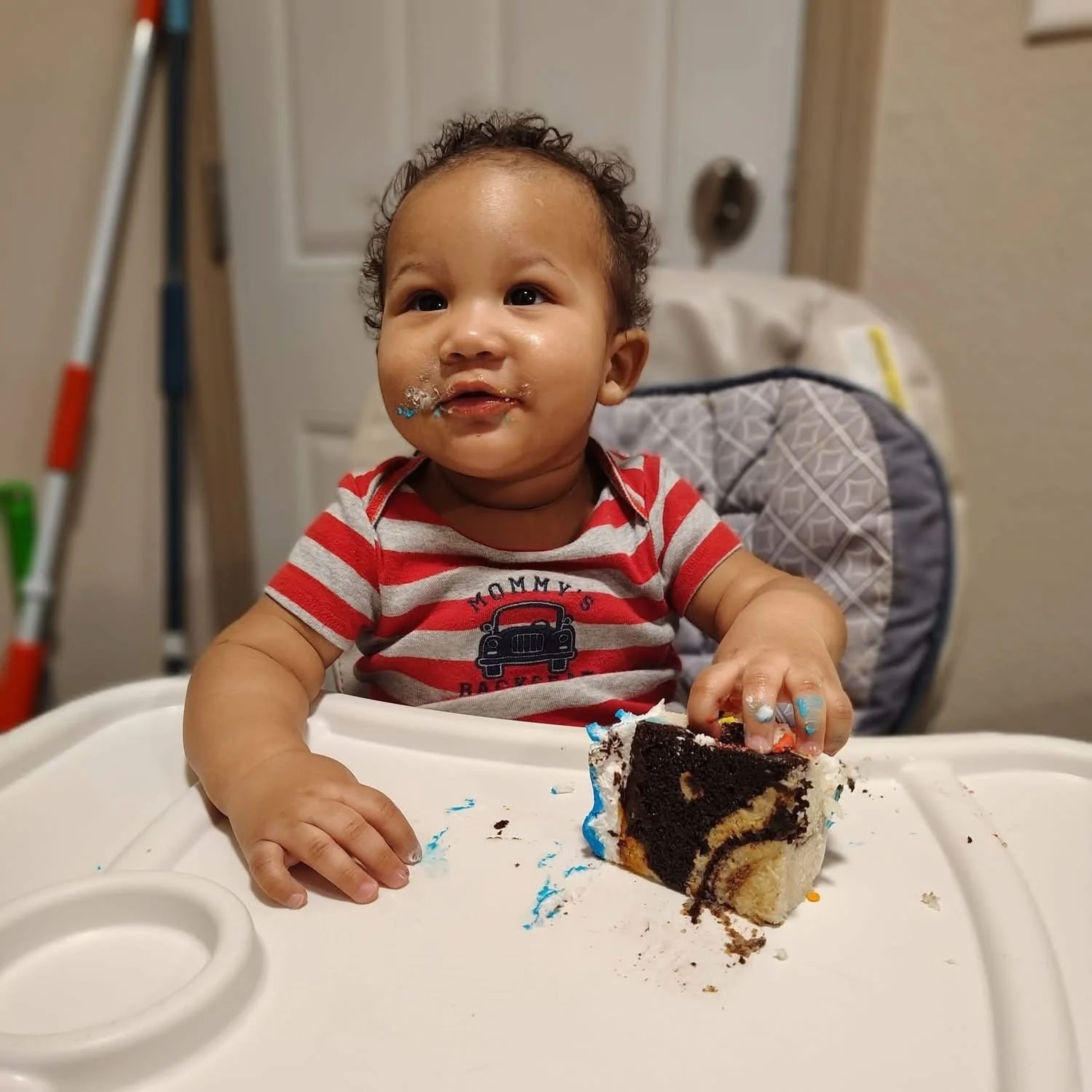 A toddler with curly hair and a striped shirt sitting in a high chair, holding a slice of cake with chocolate and vanilla layers, with some frosting and crumbs on the tray.