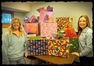 Two women standing next to a table with numerous wrapped gift boxes in colorful paper.