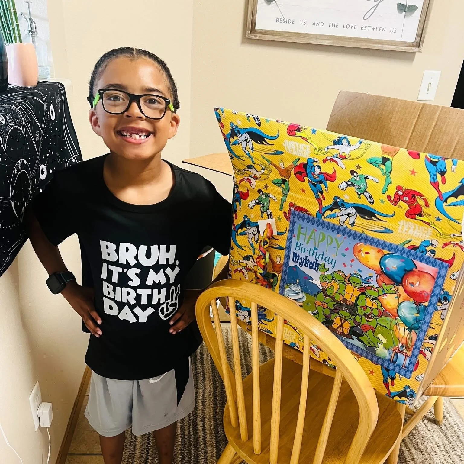A young boy with glasses smiling at a birthday party, wearing a black T-shirt that says "BRUH. IT'S MY BIRTHDAY". He stands next to a table with a large Teenage Mutant Ninja Turtles-themed birthday card and presents.