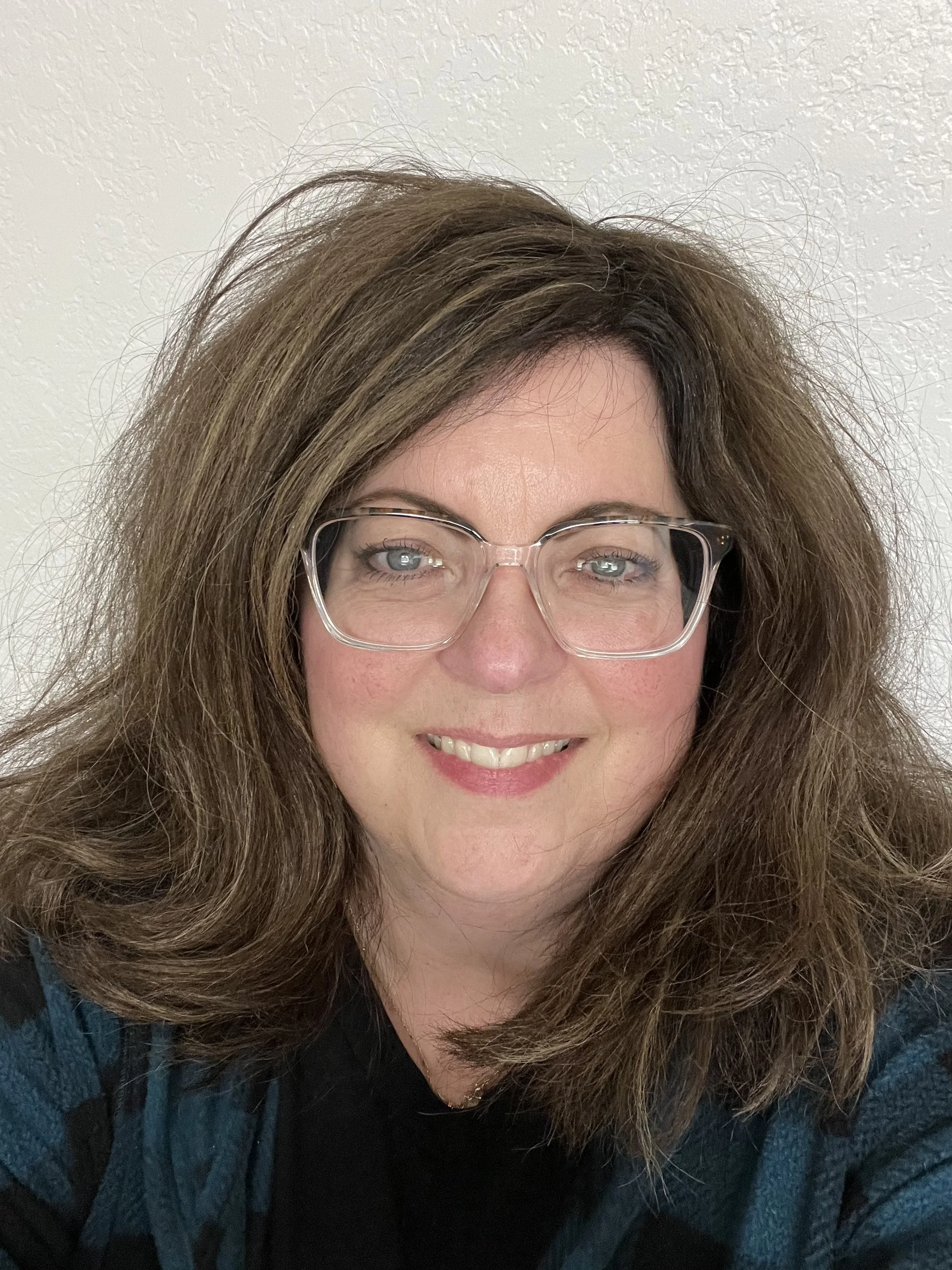 Close-up of a woman with shoulder-length brown hair, wearing glasses, smiling, against a plain light-colored wall.
