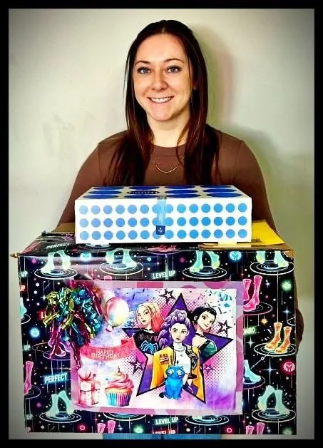 A woman standing behind a large decorated box with a colorful outer design and a smaller blue and white box on top, smiling.