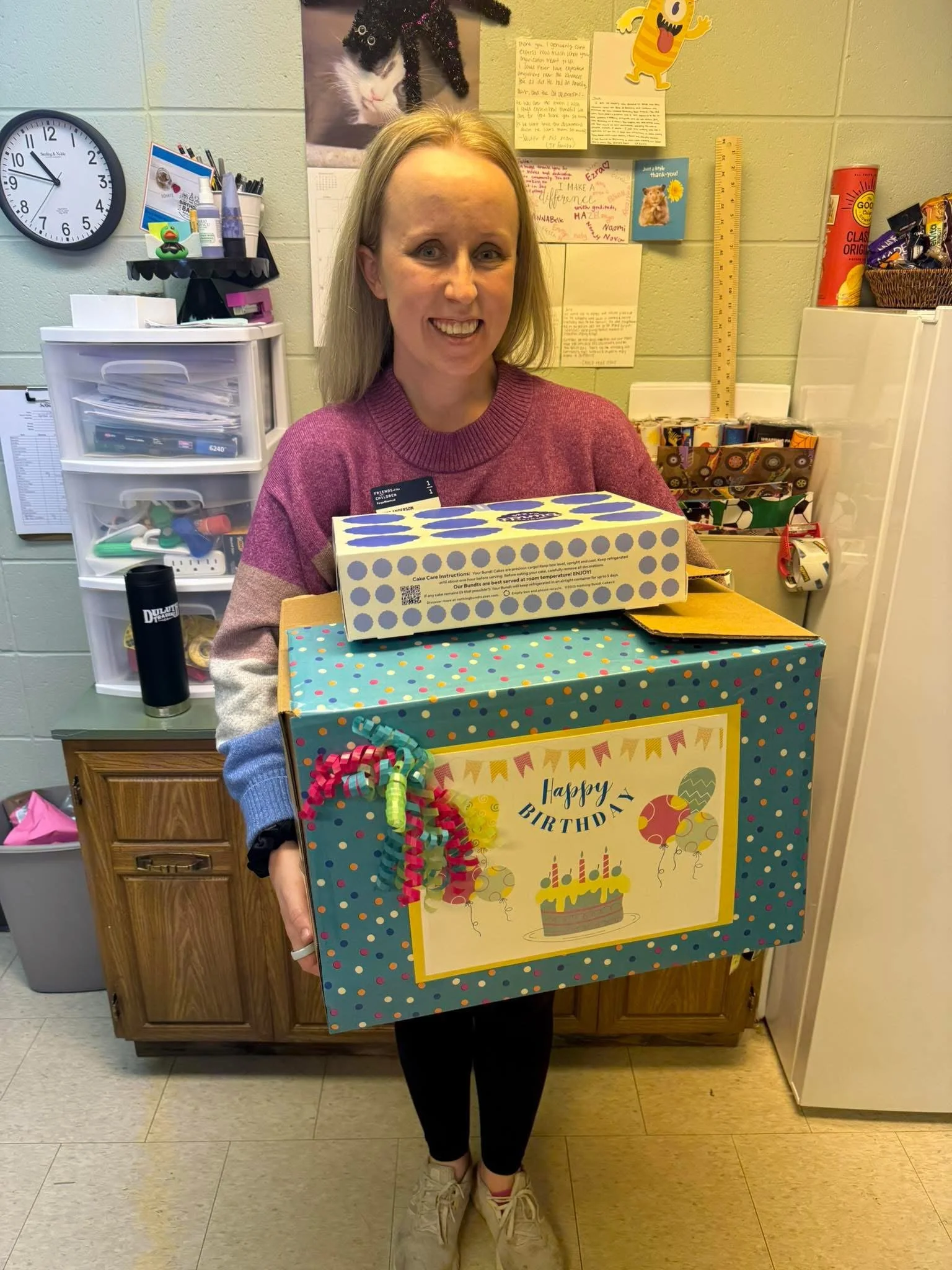 A woman with blonde hair smiling and holding large colorful birthday gift boxes in a room decorated with notes, pictures, and a cat poster on the wall.
