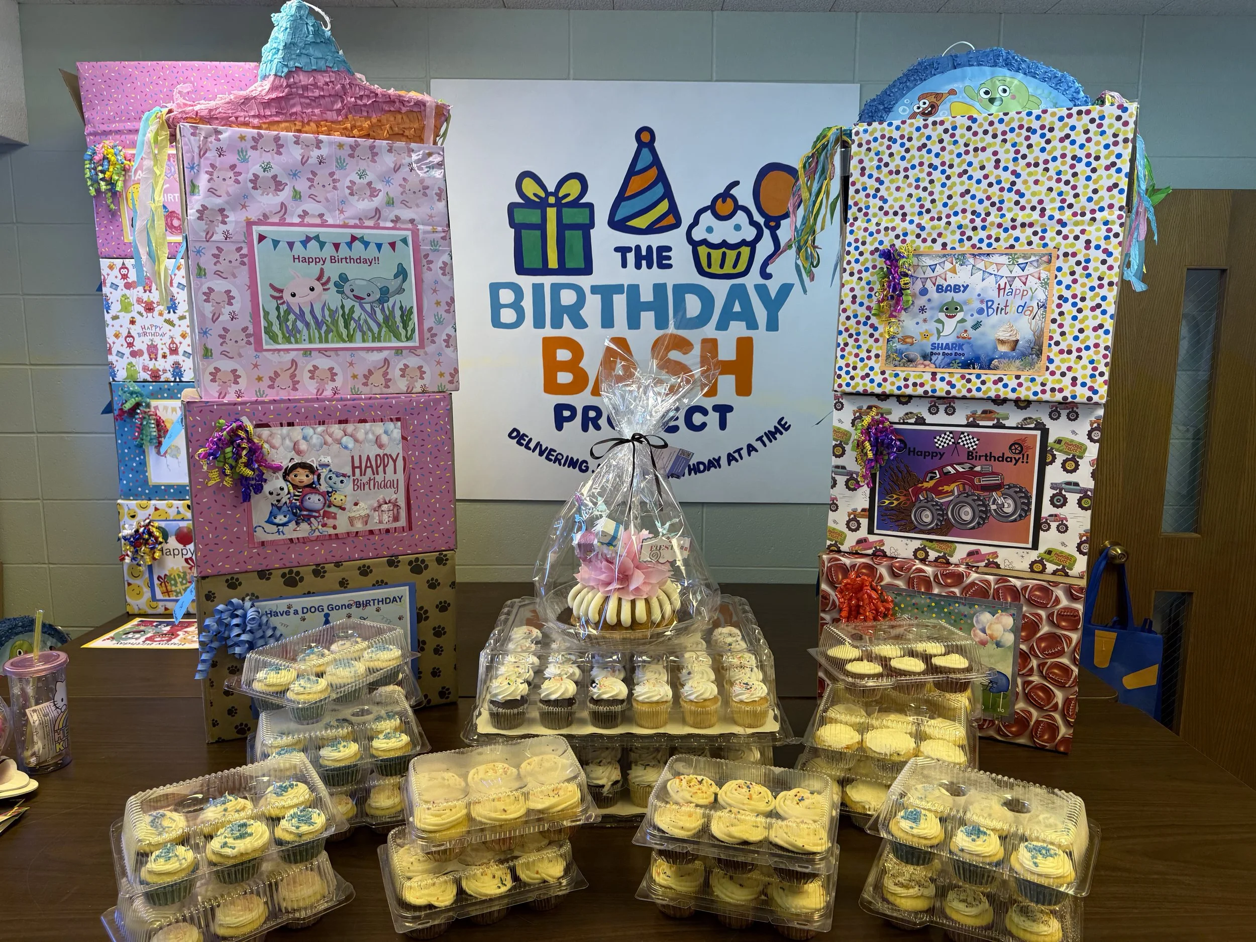 A birthday party display with cupcakes and decorated boxes featuring various cartoon characters. A large sign behind reads 'The Birthday Bash Project' with a gift, balloon, cupcake, and party hat icons.