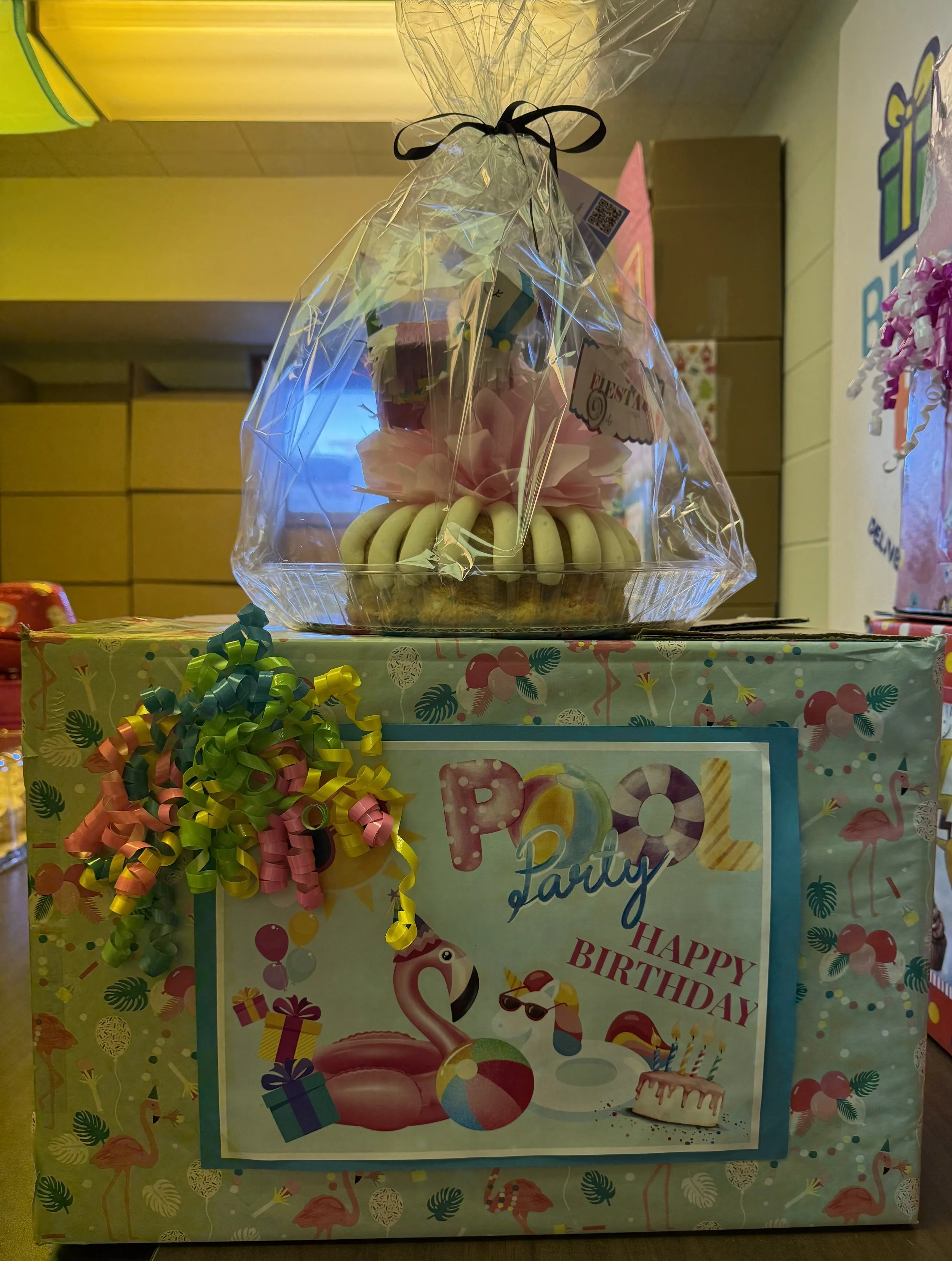 Party gift box decorated with a flamingo, balloons, and presents, with a birthday cake on top, wrapped in clear cellophane with a black ribbon.