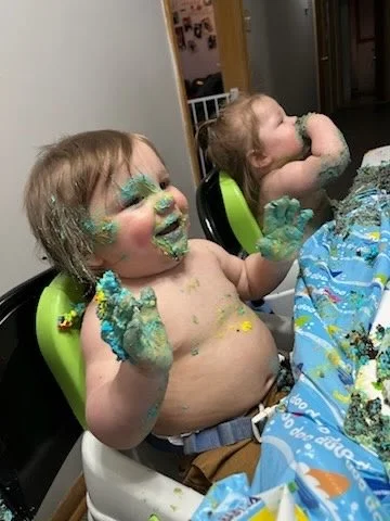 Two young children at a birthday party, covered in cake and icing, sitting at a table with a cake and colorful tablecloth.