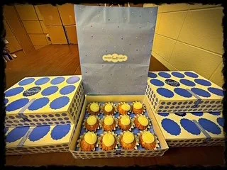 A tray of cupcakes with yellow frosting and yellow decorations, placed in front of a blue and white polka-dot patterned set of cushions or boxes, with a grey backdrop featuring a decorative cloud cutout.