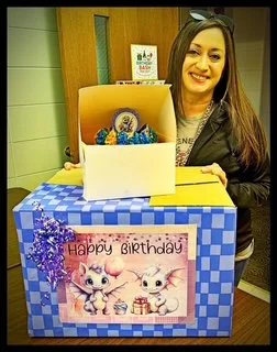 A woman with long dark hair and a leather jacket smiling next to a decorated birthday cake with a pink 'Happy Birthday' sign featuring cartoon cats, inside a blue and white checkered box with a yellow top.