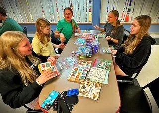 Group of five girls sitting around a table playing with colorful craft supplies and stickers in a bright room.