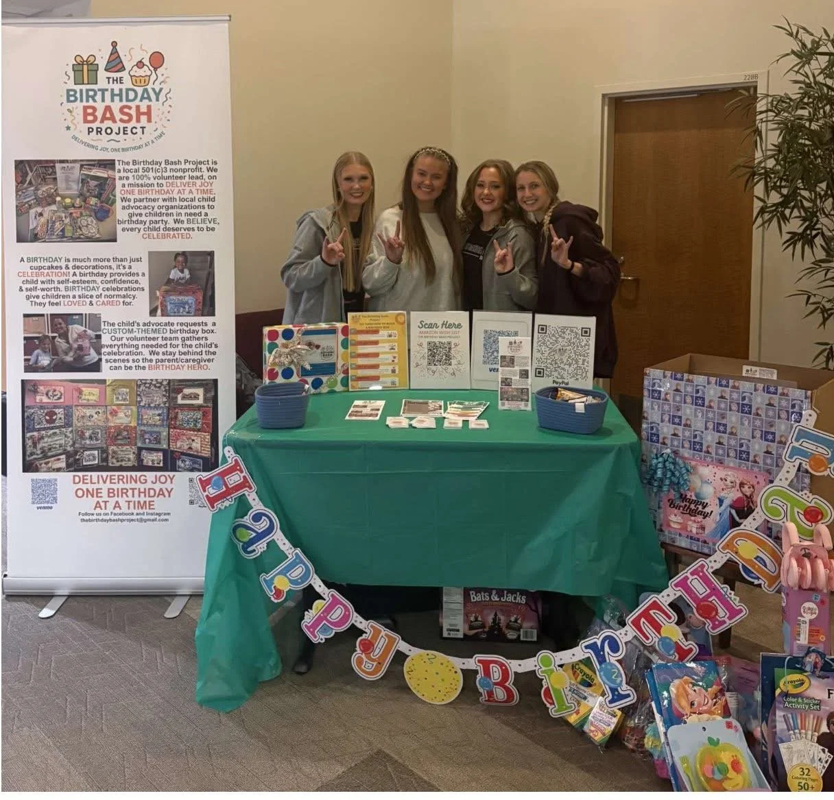 Group of four young girls standing behind a table at a birthday fundraiser event, smiling and making peace signs, with a large birthday-themed banner, informational posters, pamphlets, and gift bags on display.