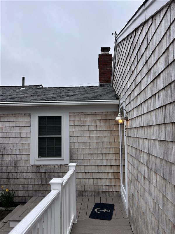 Small wooden porch with white railing outside a house with beige shingle siding, a window, and an outdoor wall-mounted light fixture over a black doormat. Overcast sky in the background.
