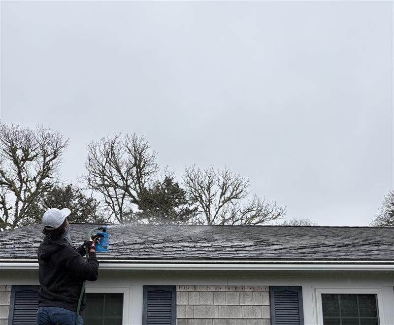 Person wearing a white cap and black jacket holding a spray bottle, cleaning the roof of a house with gray shingles and white siding, with leafless trees in the background under a cloudy sky.