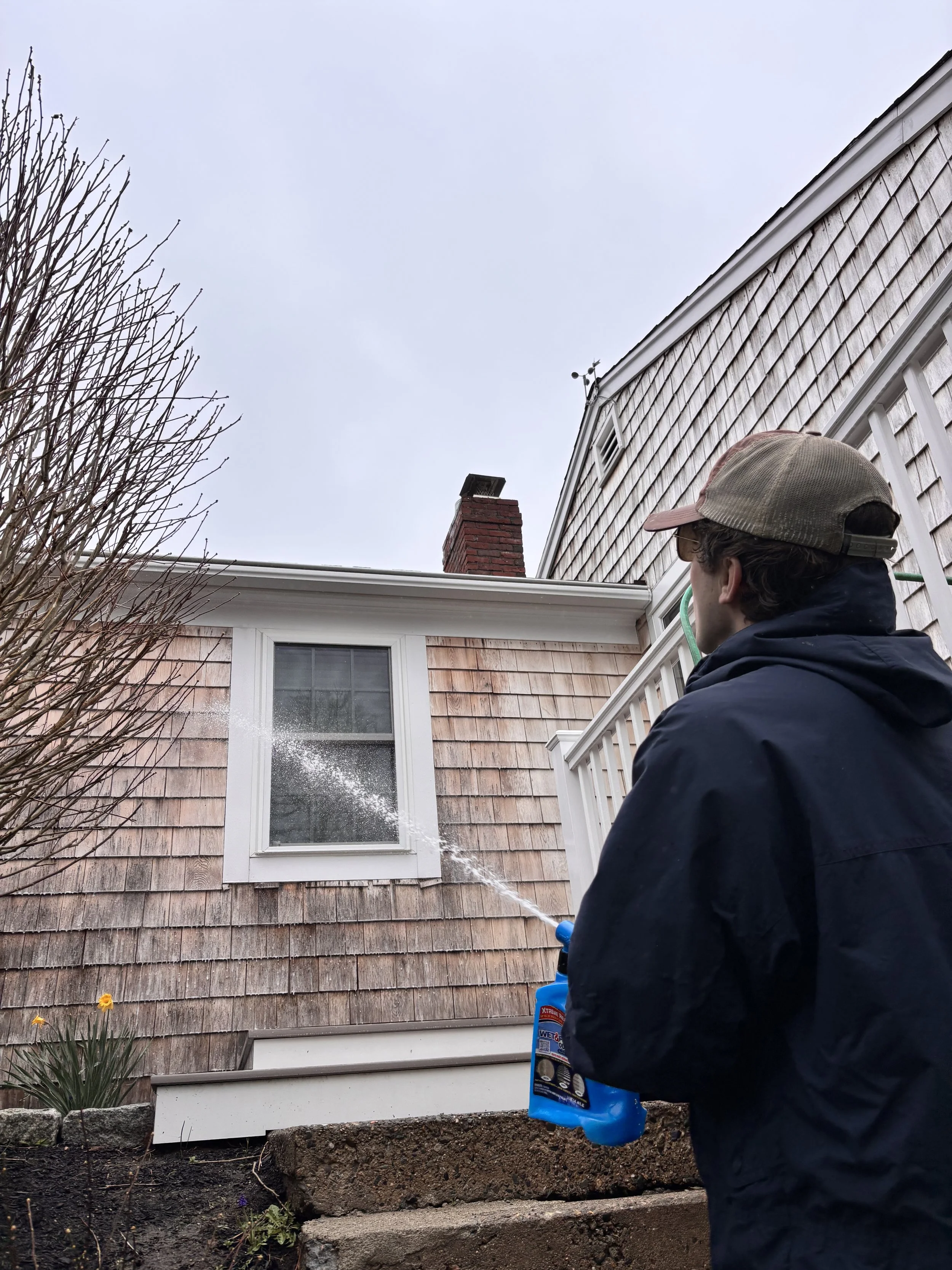A person wearing a cap and dark jacket is spraying insecticide or pesticide in the air outside a house with shingle siding, near a window, on a cloudy day.
