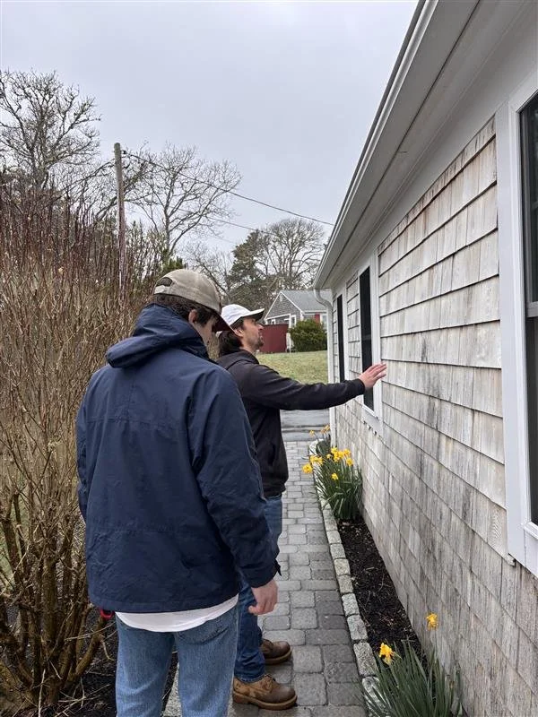 Two men are standing outside along a paved walkway next to a house with shingle siding. One is pointing at the house while the other looks on. There are yellow daffodils planted along the base of the house.