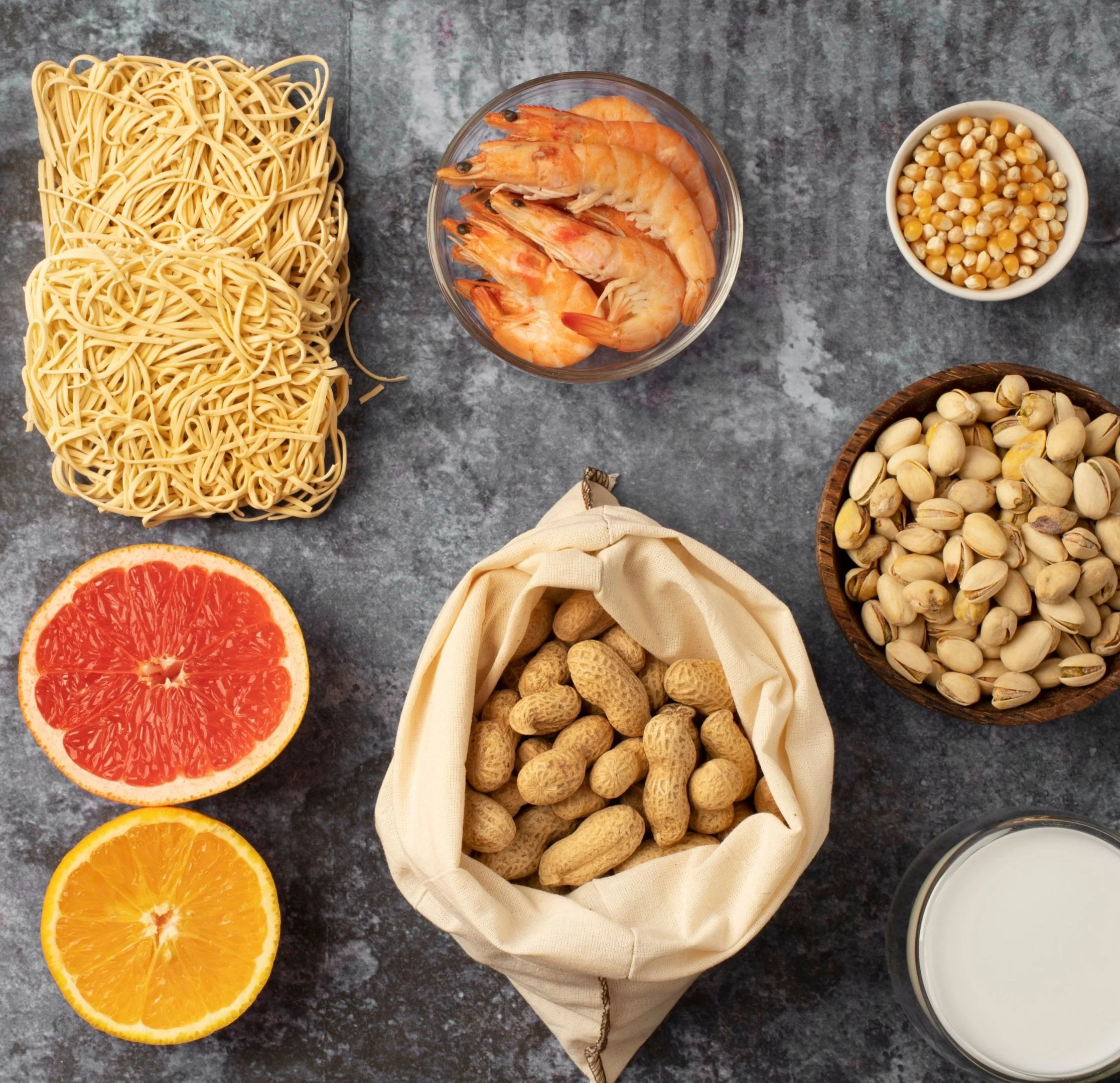 An assortment of allergen-friendly food ingredients laid out on a dark background, including gluten-free pasta, shrimp, corn, peanuts, and a glass of milk. The selection highlights a variety of protein and fiber-rich options suitable for diets.