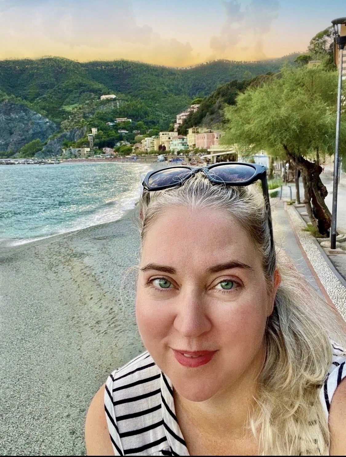 A woman with blonde hair and sunglasses on her head, taking a selfie near the beach during sunset, with colorful houses and green mountains in the background.