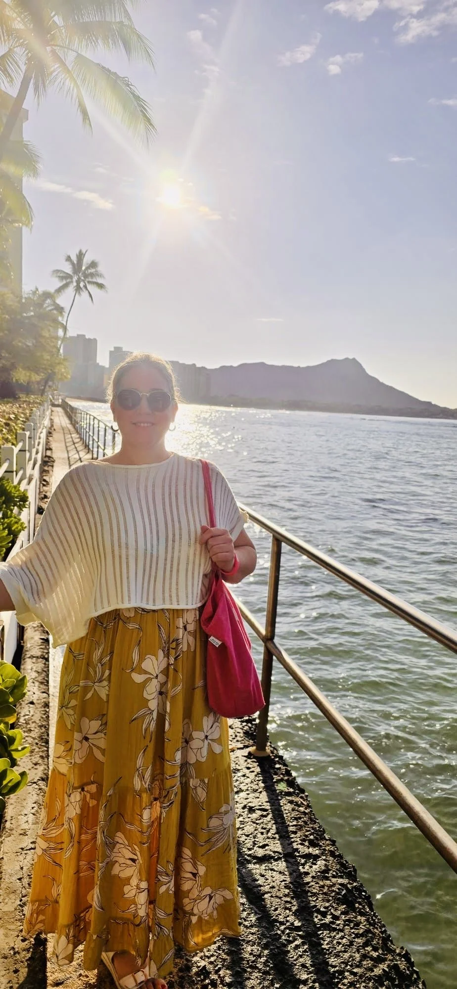 A woman with sunglasses wearing a striped top and a floral yellow skirt, standing on a pier by the water with a mountain in the background and the sun shining in the sky.