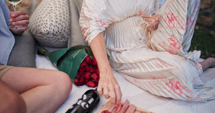 People sitting outdoors on a blanket, with one person in a flowy, floral dress, holding a drink, and next to a bouquet of red roses.