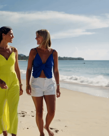Two women walking and talking on a sandy beach with the ocean in the background, under a partly cloudy sky.