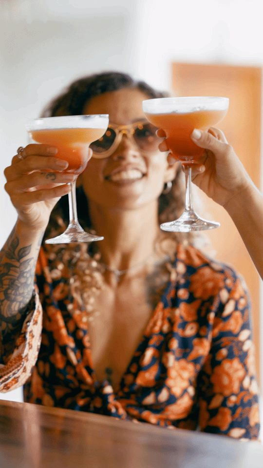A woman with curly hair and sunglasses holding up two cocktails with frothy tops, smiling at the camera.