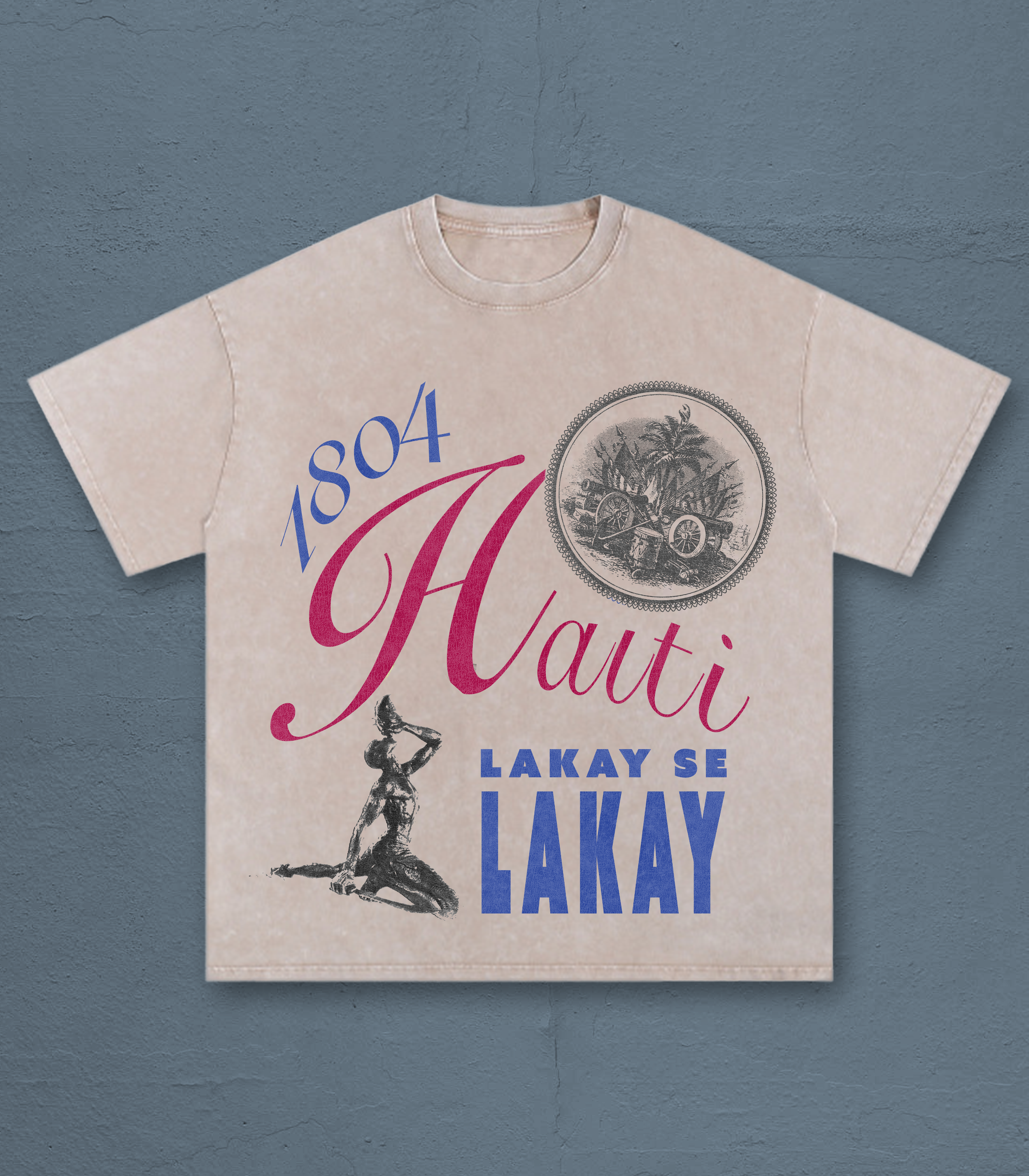 A beige T-shirt with graphic text and images, laid flat on a gray concrete surface. The shirt has a design including a vintage cannon and the phrase 'Lakay Se LaKay.'