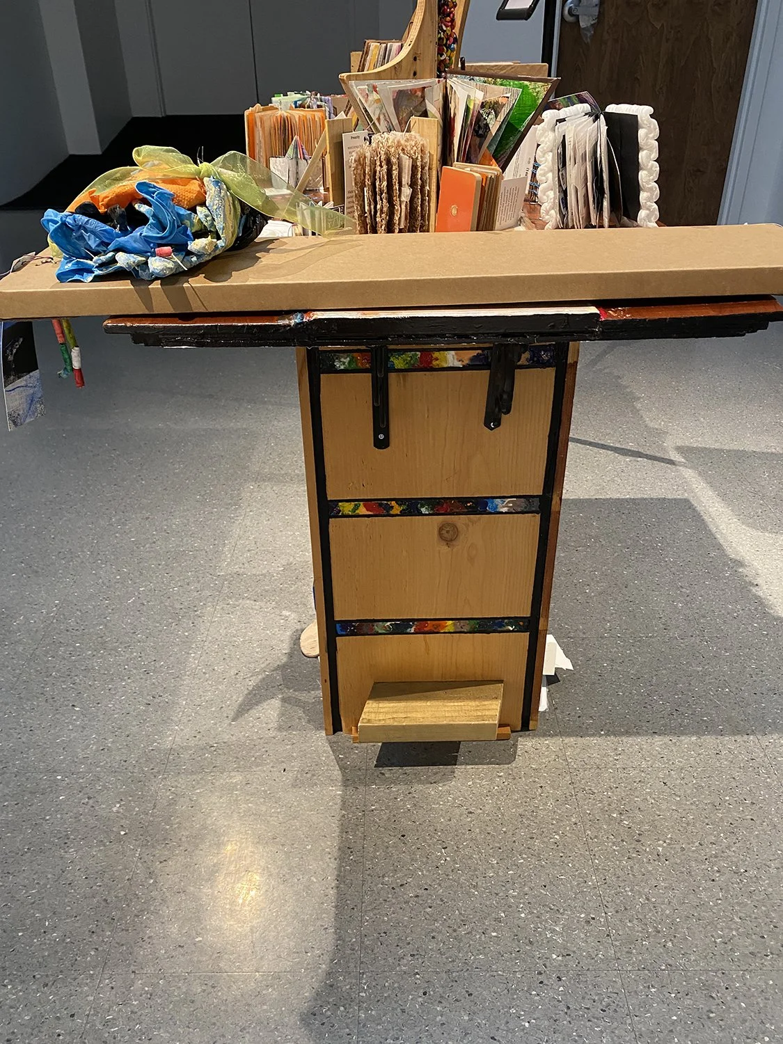 A table with a wooden base, colorful crayon inlay, and black metal supports, covered with an assortment of artist books, in a gallery.