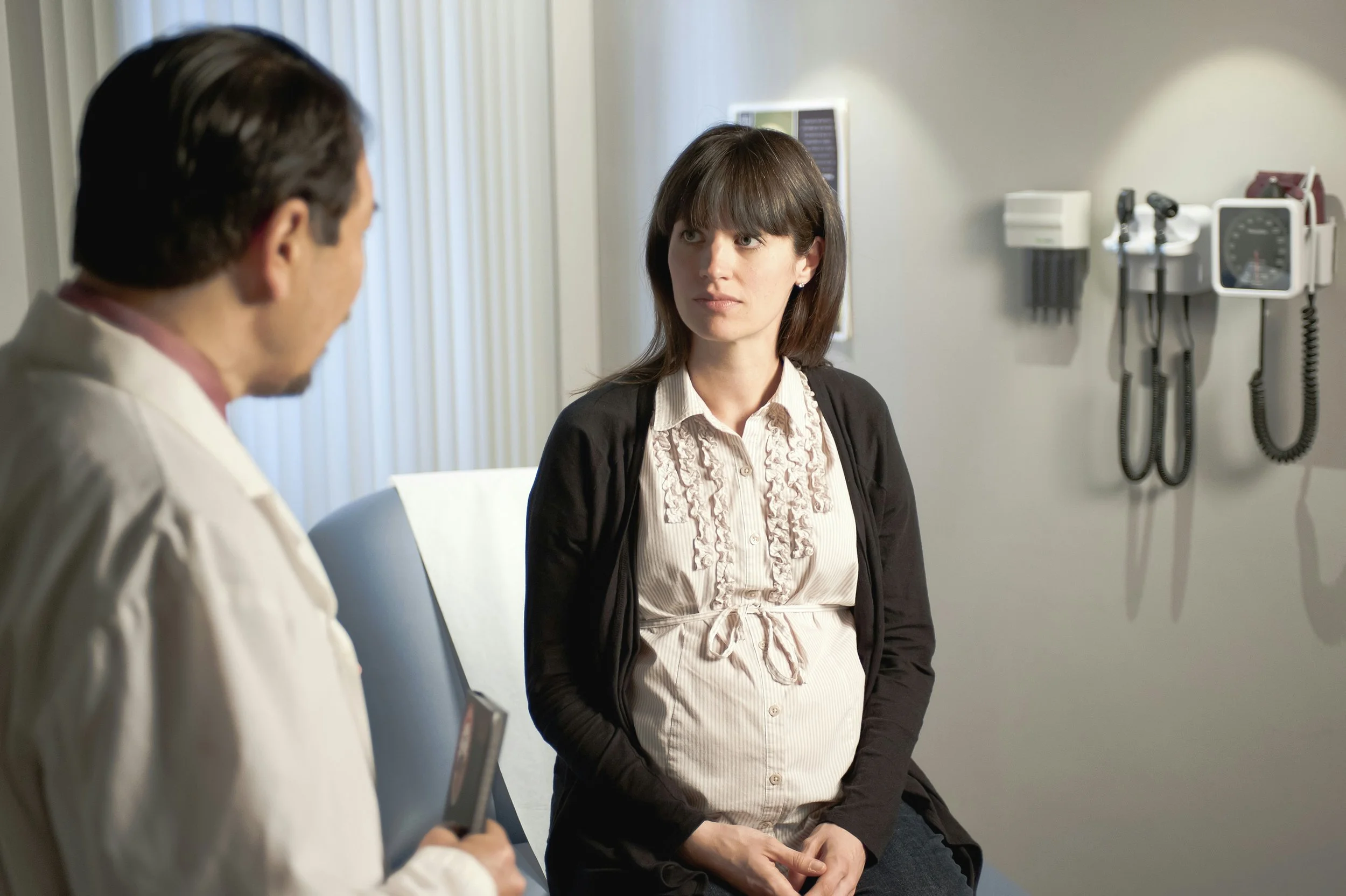 A woman sitting on a medical exam table listening attentively to a doctor in a white coat holding a clipboard, in a clinical consultation room with medical equipment mounted on the wall.