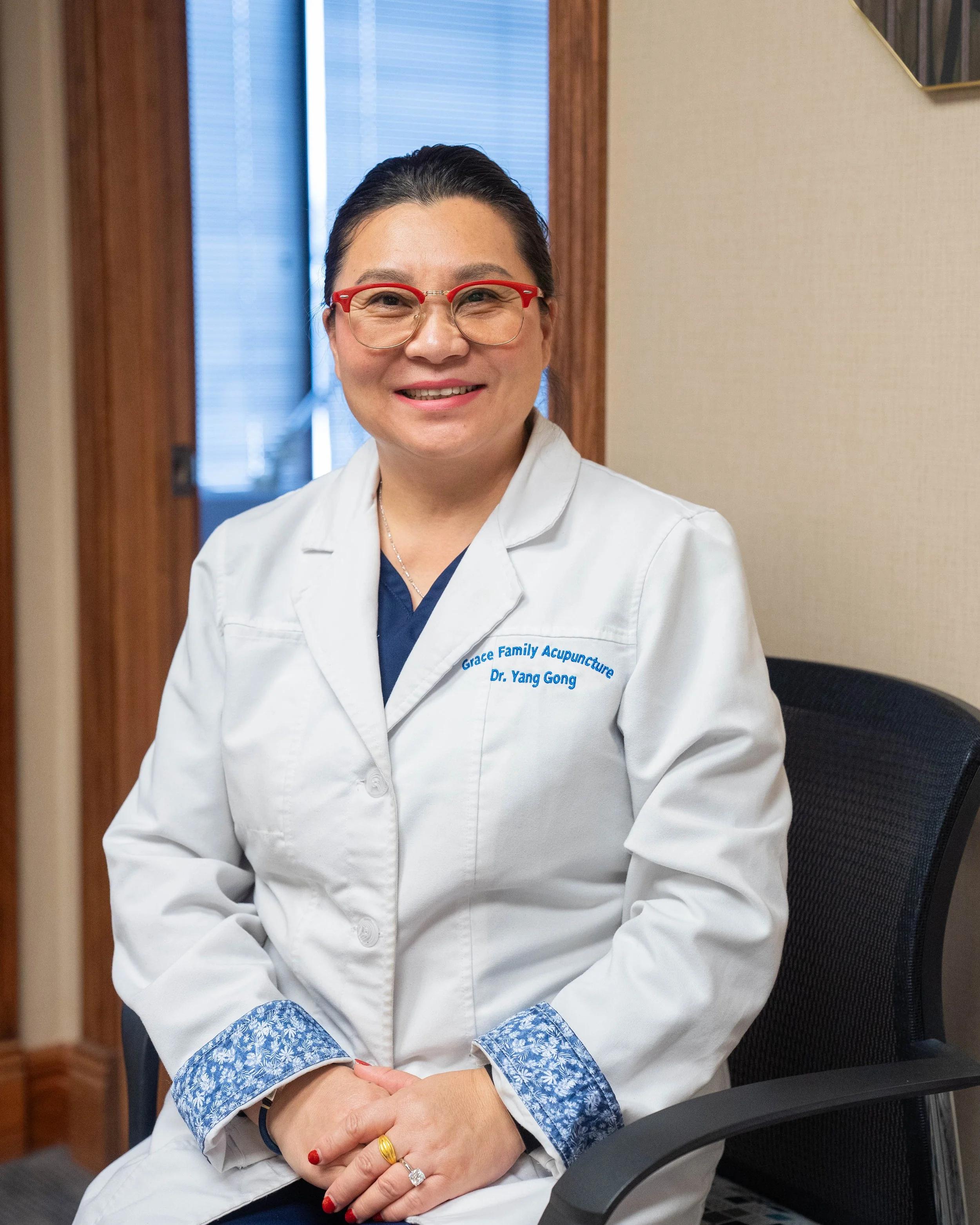A smiling woman in a white medical coat with red-rimmed glasses and rings on her fingers, sitting in an office or clinic setting with blue blinds and beige walls.