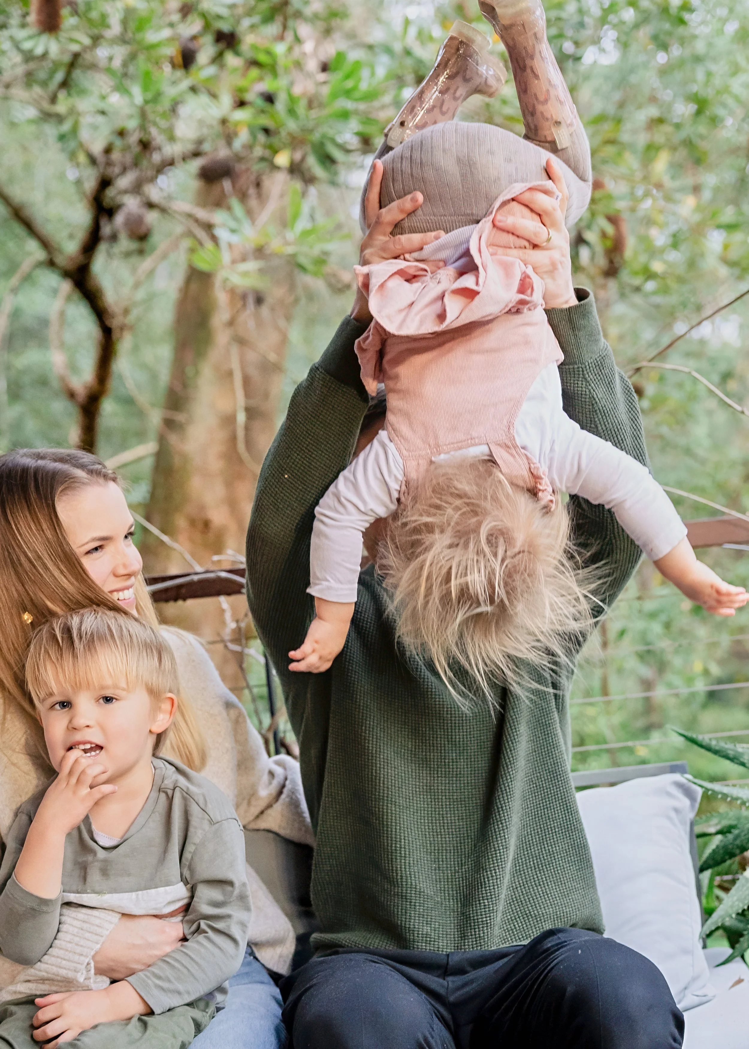 Father holding baby upside down while siblings watch and laugh during candid family photography session in Sydney