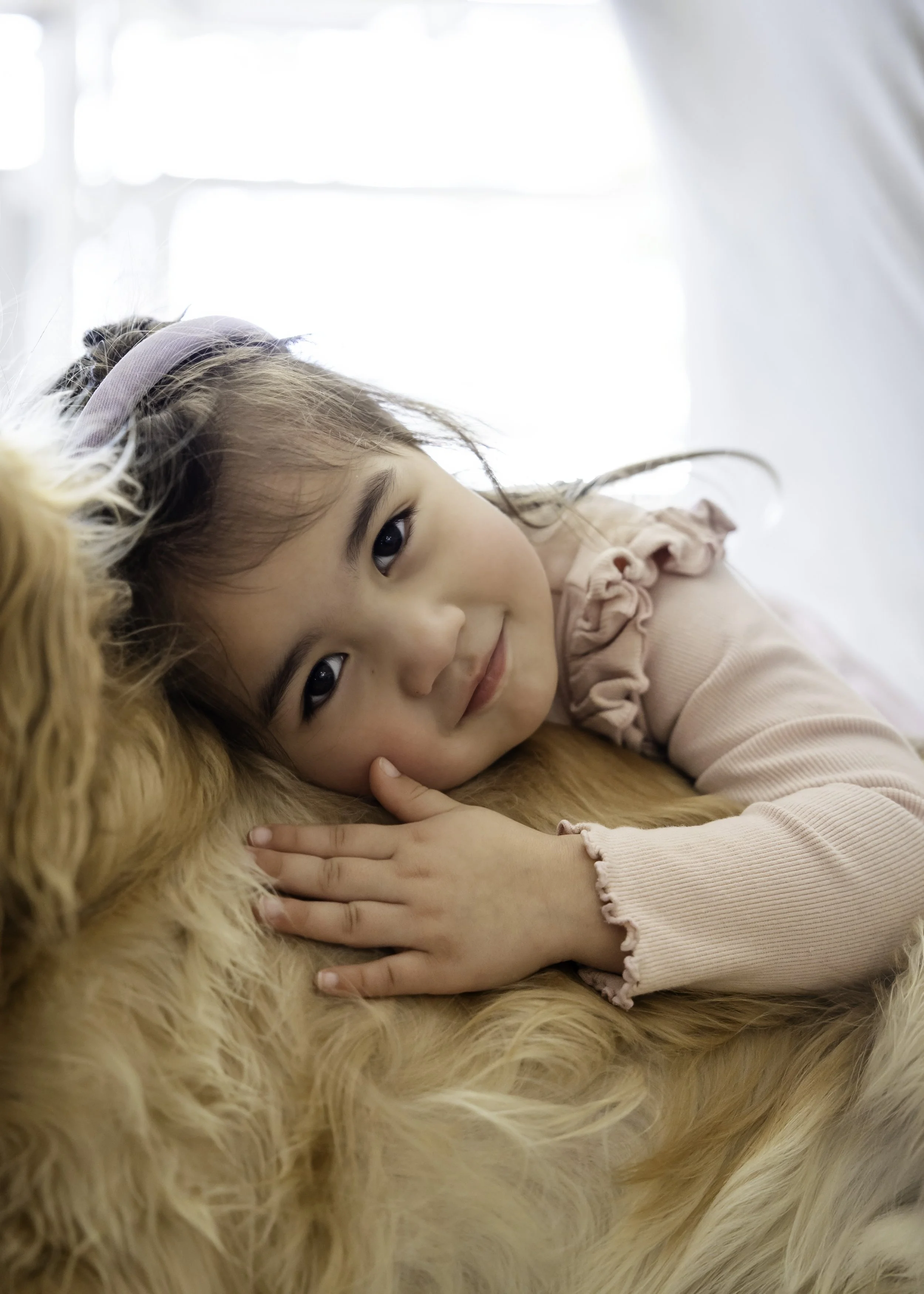 Close-up portrait of a young girl lying on a fluffy beige dog, resting her head against it during a lifestyle family photography session, soft white background.