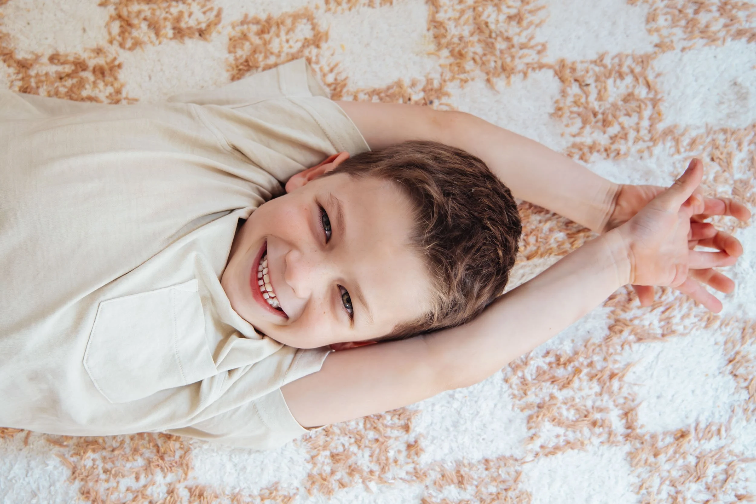A smiling young boy lying on his back on a multicolored rug, stretching his arms over his head with hands clasped, wearing a beige T-shirt.