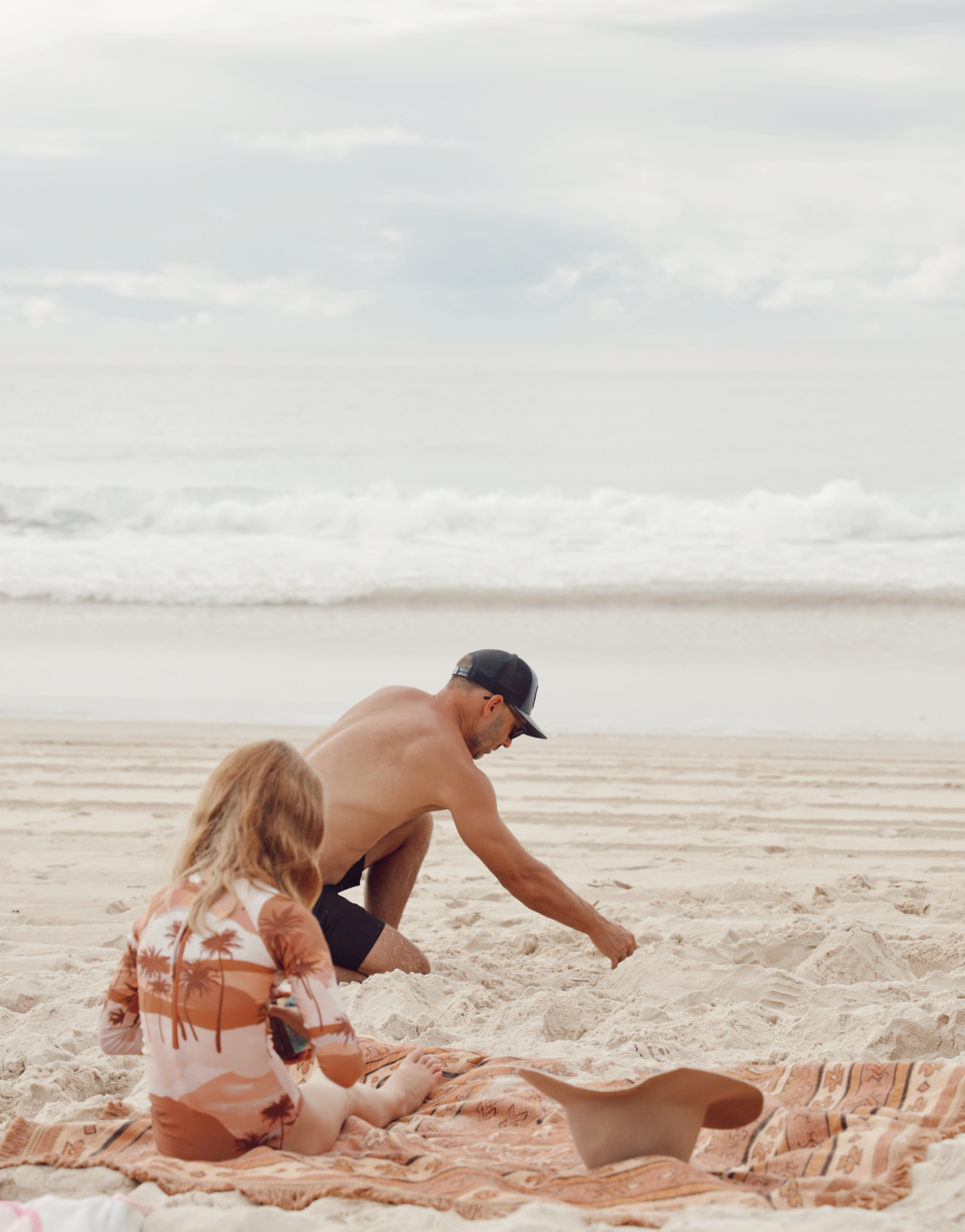 A man and a young girl on the beach, building a sandcastle on a blanket with ocean waves in the background.