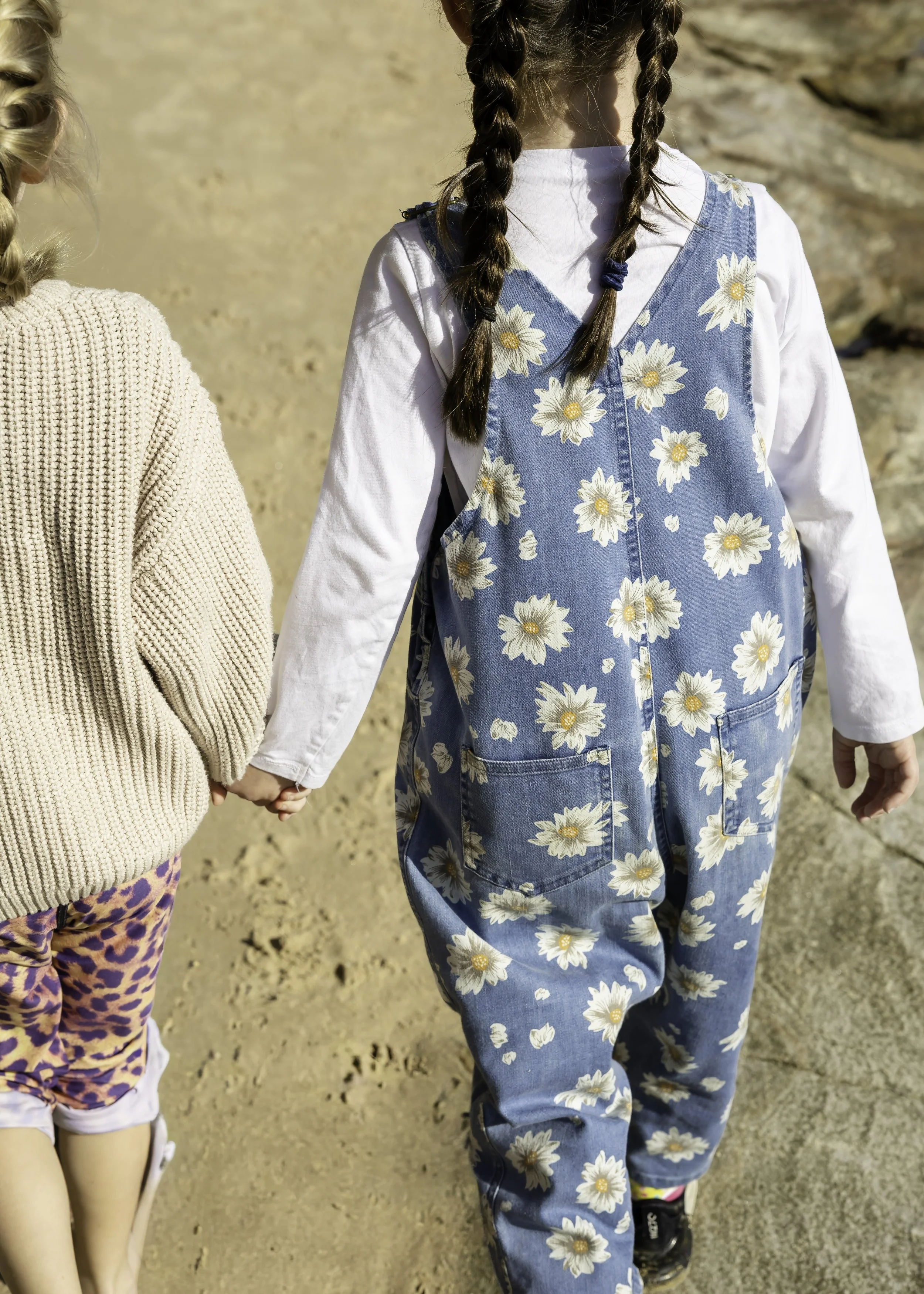 Two young girls holding hands and walking outdoors during a lifestyle family photography session, one in a beige knitted sweater, the other in a denim daisy-print dress.
