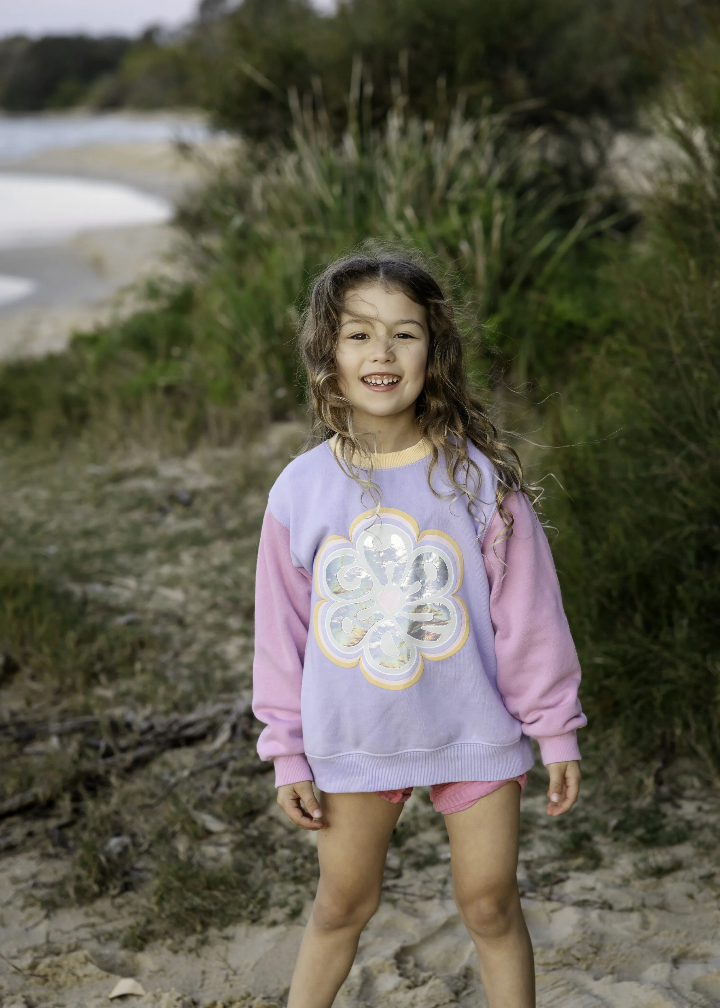 Young girl with curly brown hair smiling at the camera outdoors during a natural light family portrait session, surrounded by sandy, grassy terrain