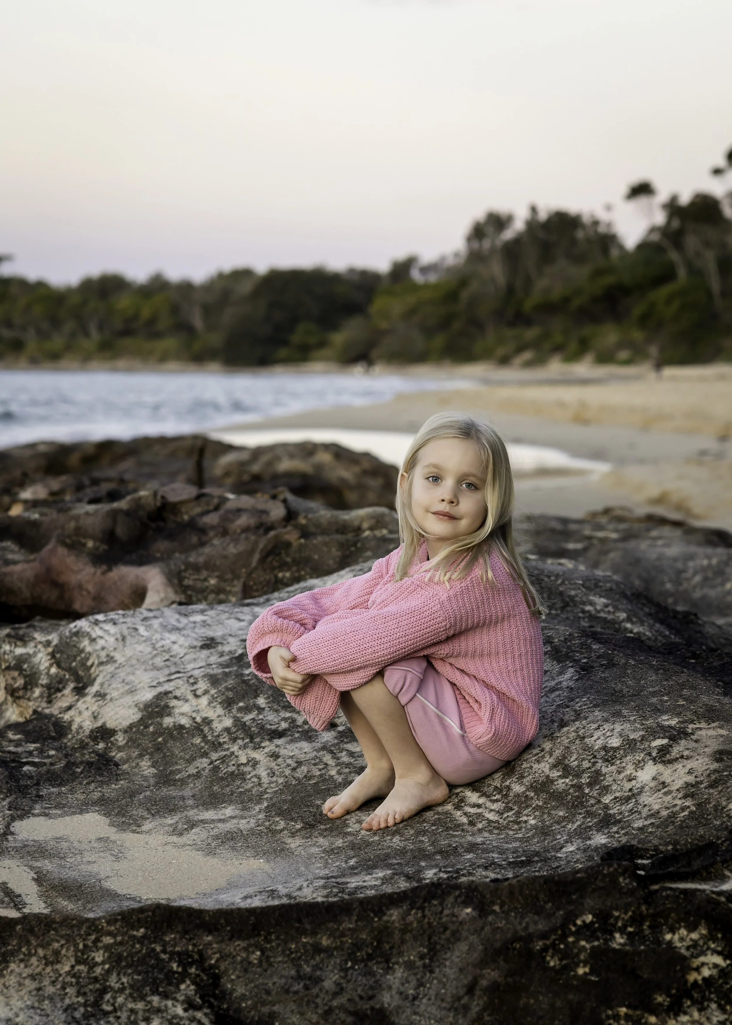 Young girl in a pink knitted sweater sitting on a rock during a beach family photography session, with sandy shore, ocean waves and green trees in the background.