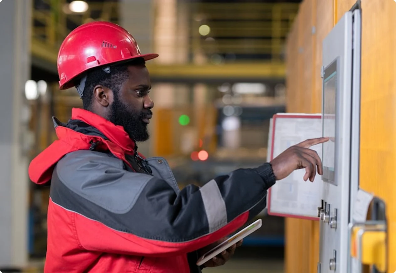 A man wearing a red safety helmet and a red and gray jacket using a touchscreen panel in a warehouse.