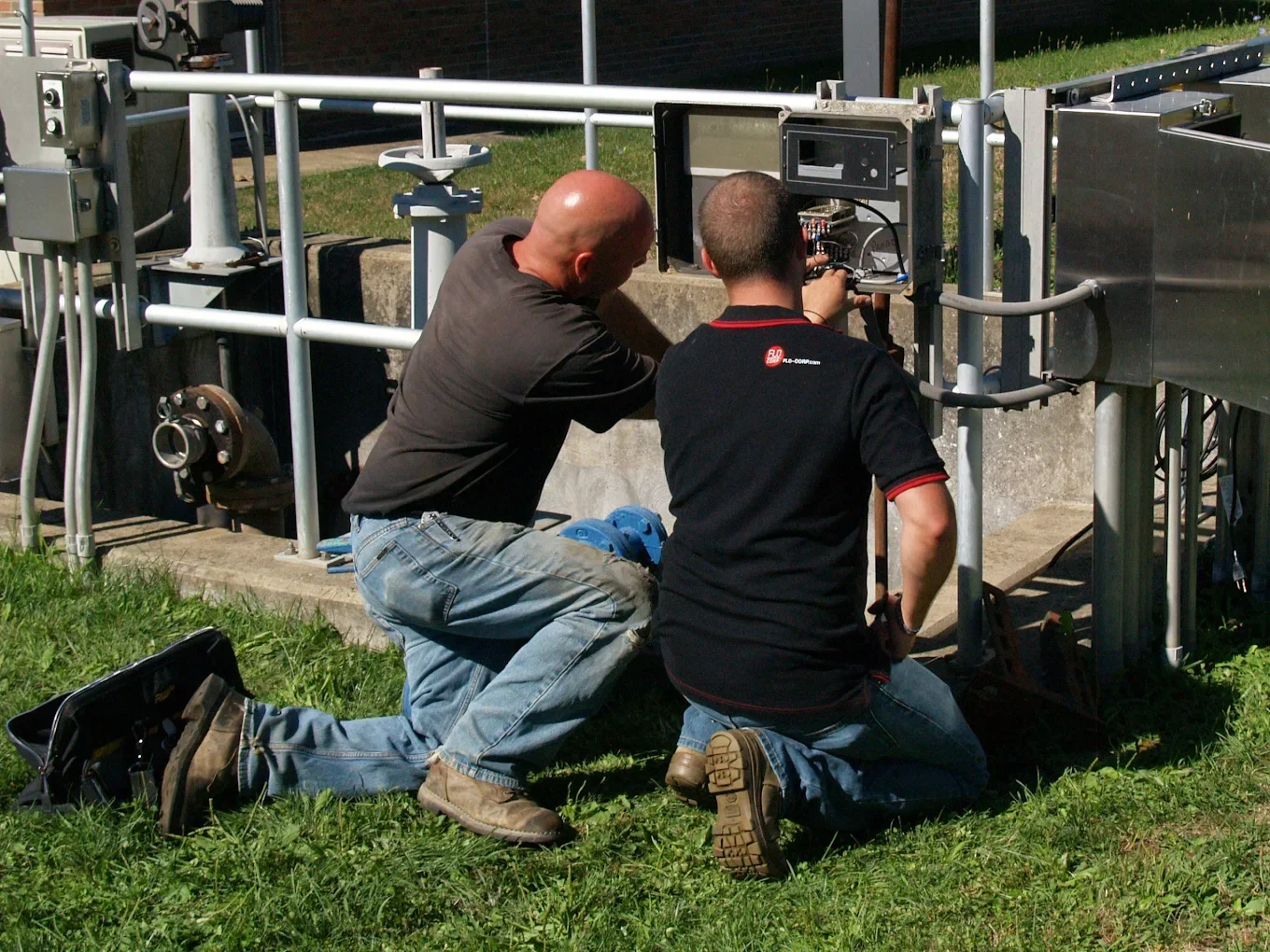 Two men working on an outdoor electrical or communication panel, with tools and equipment nearby.